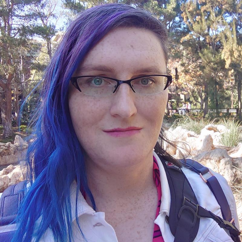 Selfie of Jennifer, taken outdoors. She's a white woman with glasses and blue-purple hair.